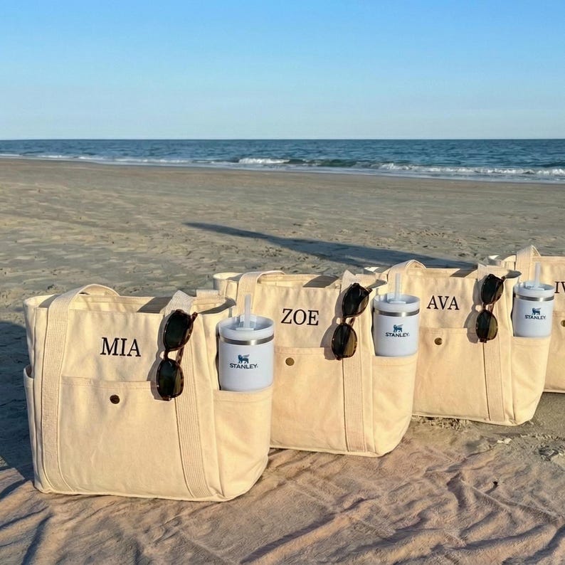May include: Beige canvas tote bags with personalized names embroidered in black. Each bag has a side pocket, sunglasses, and a light blue Stanley tumbler. The bags are on a sandy beach with the ocean in the background.