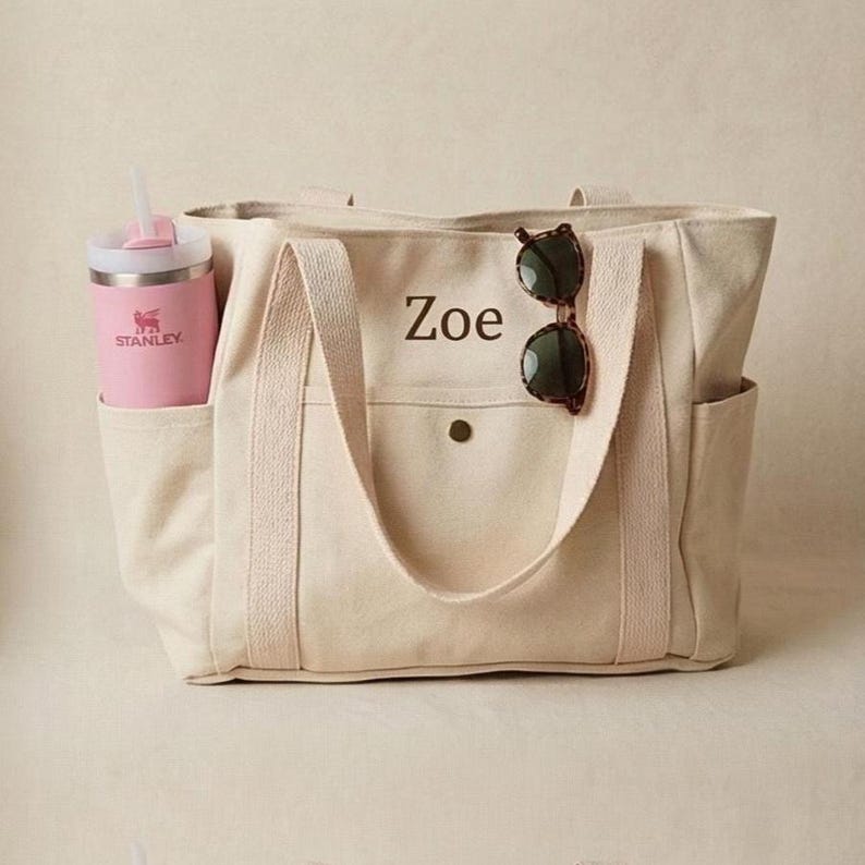 Op de afbeelding: Cr&egrave;mekleurige canvas draagtas met zij- en voorvakken. Het woord "Zoe" is op de voorkant gedrukt. Een roze Stanley waterfles met een rietje en zonnebril zitten in de zakken. De tas heeft twee handvatten.