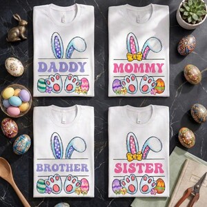 May include: Four white t-shirts with Easter designs. Each shirt has bunny ears, the words "DADDY," "MOMMY," "BROTHER," and "SISTER," paw prints, Easter eggs, and flowers. The shirts are displayed on a dark surface.