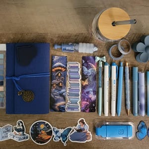 May include: An assortment of blue stationery and accessories, including a journal, pens, stickers, and washi tape. The items are arranged on a wooden surface, with a glass jar and a flower-shaped object. The color palette is primarily shades of blue.