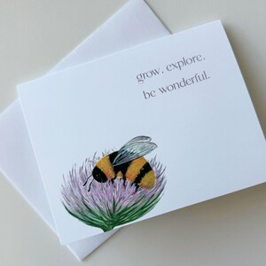 Pode incluir: Cartão branco com uma ilustração em aquarela de um zangão em uma flor de cardo roxo. O cartão tem o texto "grow. explore. be wonderful." impresso em uma fonte simples. Um envelope branco está atrás do cartão.