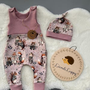Puede incluir: Un conjunto de ropa para bebé con un pelele rosa y un gorro a juego, ambos adornados con un estampado de animales del bosque. El conjunto incluye un letrero de madera con una ilustración de erizo y el texto "Liam & Luppy".
