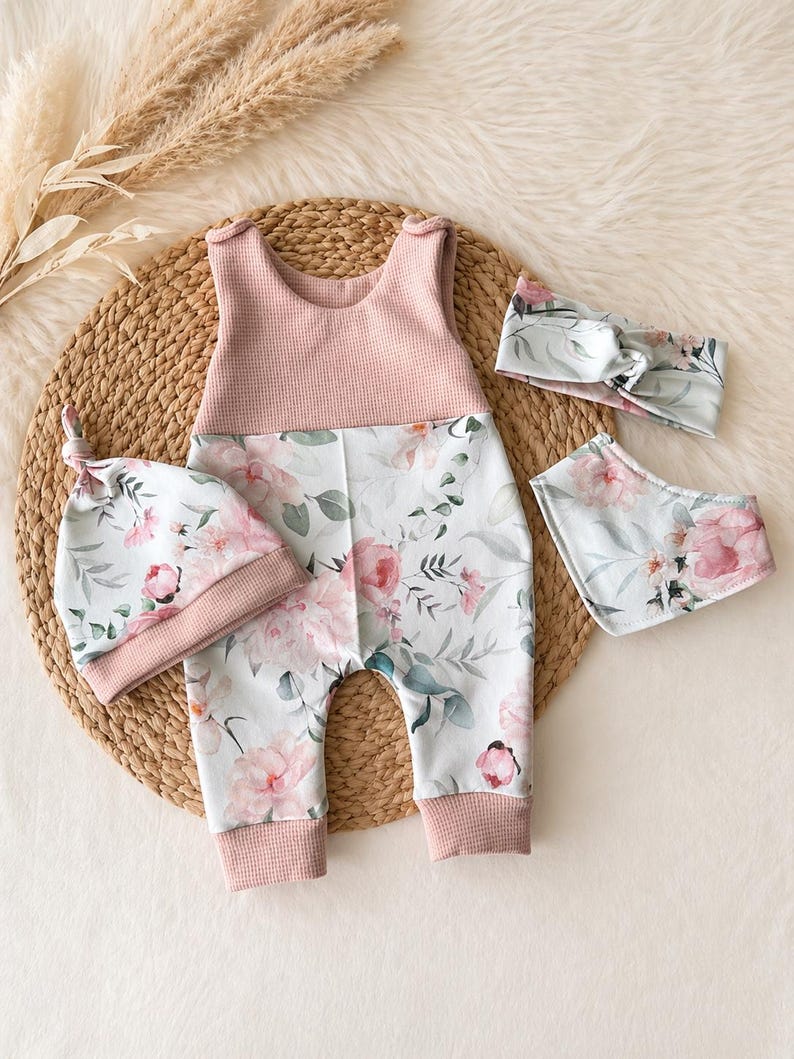 May include: A baby clothing set featuring a pink and floral print romper, a matching knotted hat, a bib, and a headband. The romper has a pink waffle-knit bodice and floral print pants. The hat, bib, and headband have a coordinating floral pattern.
