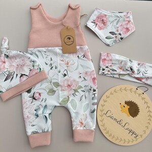 May include: A baby clothing set featuring a pink and white floral print. The set includes a romper, hat, bib, and headband. The romper and hat have a pink waffle-knit top. A wooden sign with a hedgehog illustration is also included.