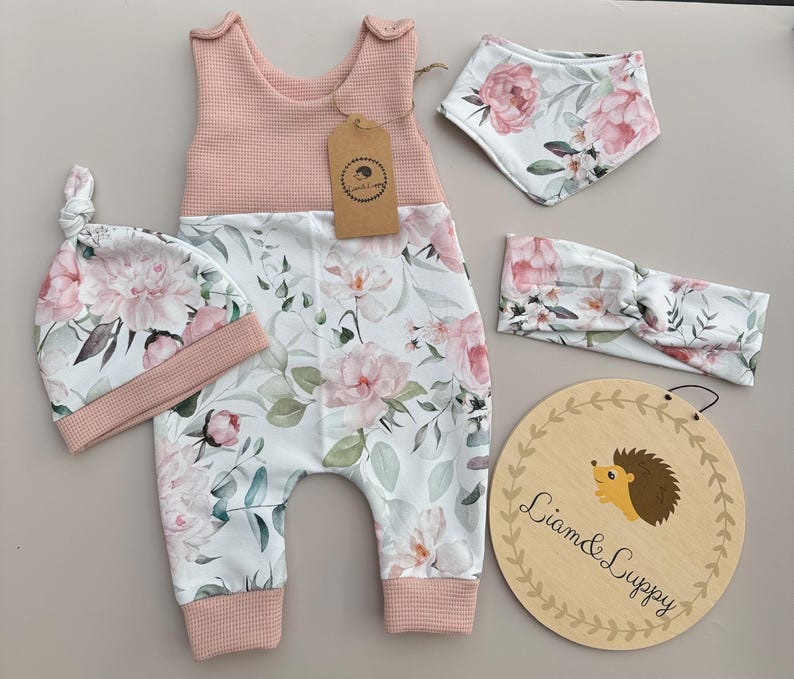 May include: A baby clothing set featuring a pink and white floral print. The set includes a romper, hat, bib, and headband. The romper and hat have a pink waffle-knit top. A wooden sign with a hedgehog illustration is also included.