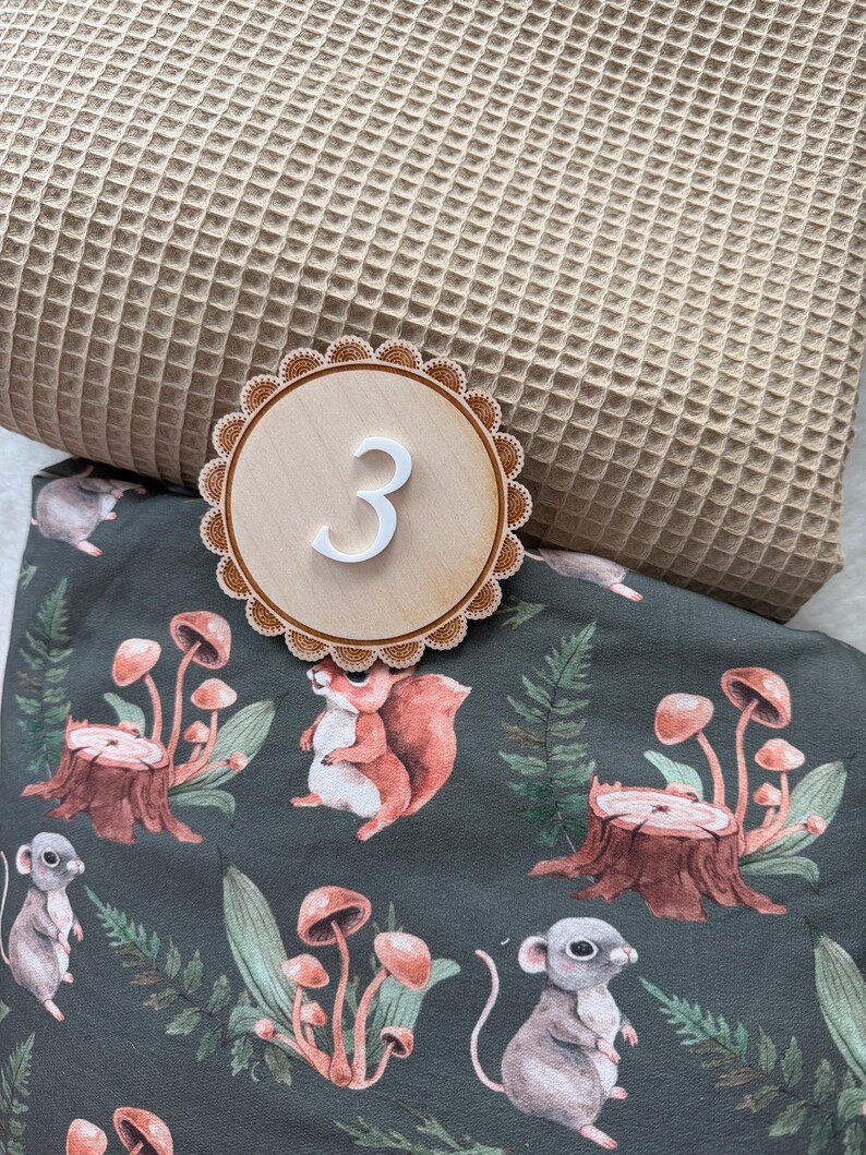 Puede incluir: Un n&uacute;mero tres de madera con un numeral blanco, sobre un borde festoneado, se asienta sobre una tela verde oscuro con un estampado de bosque de ardillas, setas y ratones. Una tela de tejido de gofre beige est&aacute; en el fondo.