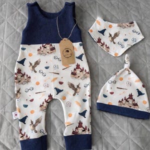 Puede incluir: Un conjunto de bebé de tres piezas con una combinación de colores azul marino y blanco. El conjunto incluye un mono, un babero y un gorro anudado, todos adornados con un estampado caprichoso de un castillo, búhos y otros elementos mágicos. El mono tiene una parte superior azul marino y una inferior estampada.
