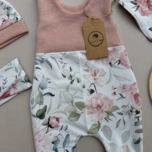 May include: A baby romper set featuring a pink waffle-knit top and floral print bottom with matching hat and bib. The floral print includes pink peonies and green foliage on a white background. A brown tag with a hedgehog logo is attached to the romper.