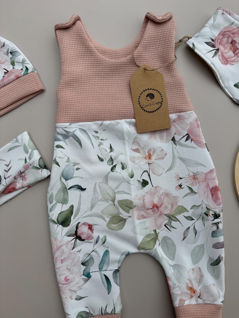 May include: A baby romper set featuring a pink waffle-knit top and floral print bottom with matching hat and bib. The floral print includes pink peonies and green foliage on a white background. A brown tag with a hedgehog logo is attached to the romper.