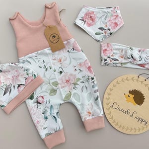 May include: A baby outfit set featuring a pink and floral print. The set includes a jumpsuit, hat, bib, and headband. A wooden sign with the text "Liam & Luppy" and a hedgehog illustration is also included.
