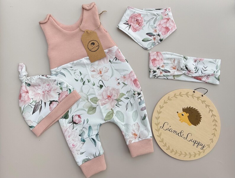 May include: A baby outfit set featuring a pink and floral print. The set includes a jumpsuit, hat, bib, and headband. A wooden sign with the text "Liam & Luppy" and a hedgehog illustration is also included.