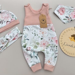 May include: A baby clothing set featuring a pink and white floral print. The set includes a jumpsuit, hat, headband, and bib. The jumpsuit has a pink waffle-knit top and floral print bottom. A wooden sign with a hedgehog illustration is also included.