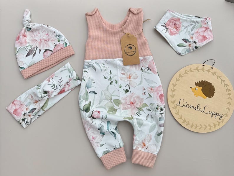 May include: A baby clothing set featuring a pink and white floral print. The set includes a jumpsuit, hat, headband, and bib. The jumpsuit has a pink waffle-knit top and floral print bottom. A wooden sign with a hedgehog illustration is also included.