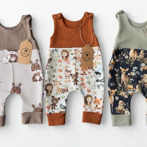 Puede incluir: Tres peleles para bebés con diferentes estampados. Uno es marrón y beige con conejos y patos, otro es naranja y blanco con animales del bosque, y el último es verde y negro con ciervos y flores.