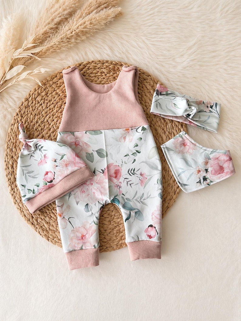 May include: A baby clothing set featuring a pink and floral print romper, a matching knotted hat, a bib, and a headband. The romper has a pink waffle-knit bodice and floral print pants. The hat, bib, and headband have a coordinating floral pattern.