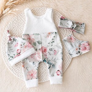 May include: A baby outfit set featuring a white waffle-knit top and floral print pants with pink flowers and green leaves. The set includes a matching hat, headband, and bib. The items are arranged on a woven mat.