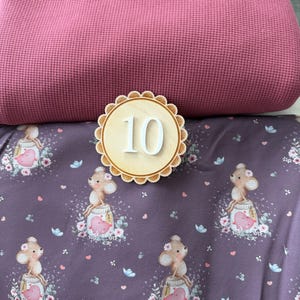 Puede incluir: Un n&uacute;mero 10 de madera con borde festoneado se encuentra sobre una tela morada estampada con ratones de dibujos animados, corazones y flores. Un tejido rosa grande y texturizado est&aacute; en el fondo.
