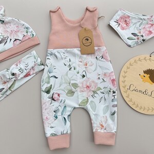 May include: A baby clothing set featuring a pink and white floral print. The set includes a romper, hat, headband, and bib. The romper has a pink waffle-knit top and the floral print on the bottom. A wooden sign with a hedgehog illustration is also included.