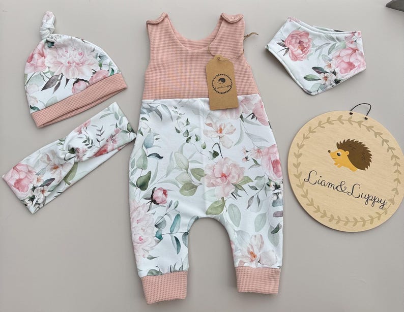 May include: A baby clothing set featuring a pink and white floral print. The set includes a romper, hat, headband, and bib. The romper has a pink waffle-knit top and the floral print on the bottom. A wooden sign with a hedgehog illustration is also included.