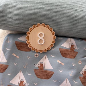 Puede incluir: Un n&uacute;mero ocho de madera con borde festoneado sobre tela azul con estampado n&aacute;utico. El estampado presenta barcos marrones con velas blancas, osos, anclas y gaviotas. Un tejido azul texturizado est&aacute; en el fondo.