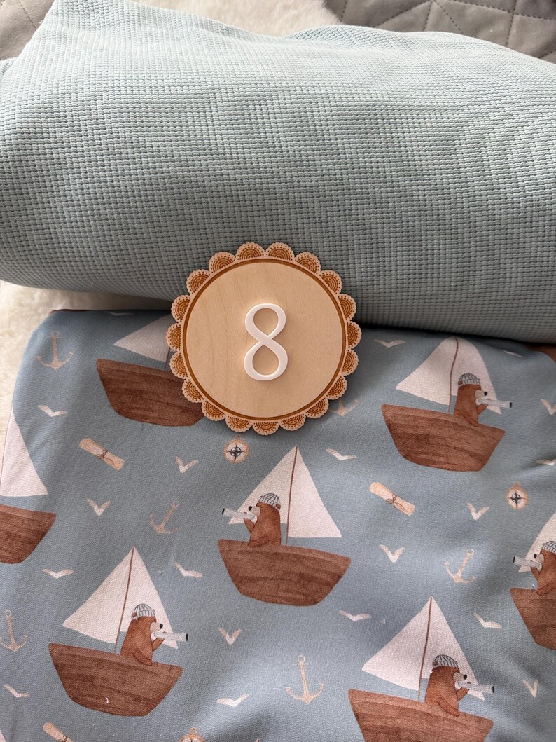 Puede incluir: Un n&uacute;mero ocho de madera con borde festoneado sobre tela azul con estampado n&aacute;utico. El estampado presenta barcos marrones con velas blancas, osos, anclas y gaviotas. Un tejido azul texturizado est&aacute; en el fondo.
