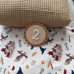 Puede incluir: Una manta de tejido de gofre beige claro se combina con un letrero redondo de madera con el n&uacute;mero "2" en blanco. Debajo, una tela blanca est&aacute; impresa con b&uacute;hos, gafas, llaves y otras ilustraciones tem&aacute;ticas m&aacute;gicas.