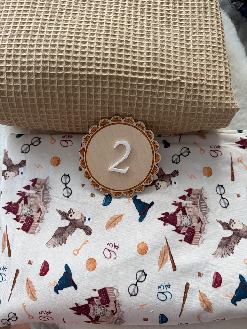 Puede incluir: Una manta de tejido de gofre beige claro se combina con un letrero redondo de madera con el n&uacute;mero "2" en blanco. Debajo, una tela blanca est&aacute; impresa con b&uacute;hos, gafas, llaves y otras ilustraciones tem&aacute;ticas m&aacute;gicas.