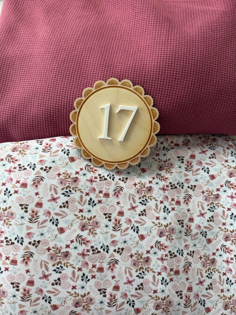 Puede incluir: Un n&uacute;mero 17 de madera, con un borde festoneado, se encuentra sobre una tela floral y una tela rosa texturizada. El n&uacute;mero est&aacute; en una fuente blanca y limpia. La tela floral tiene un fondo blanco con flores rosas y marrones.