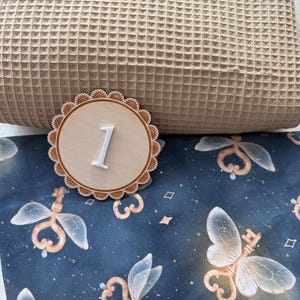 Puede incluir: Un disco de madera con el n&uacute;mero uno y un borde festoneado. El n&uacute;mero es blanco y en relieve. El disco est&aacute; sobre una tela azul con un patr&oacute;n de llaves de hadas y una almohada beige gofrada.