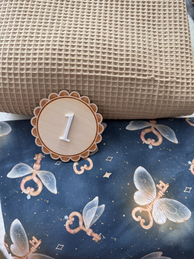 Puede incluir: Un disco de madera con el n&uacute;mero uno y un borde festoneado. El n&uacute;mero es blanco y en relieve. El disco est&aacute; sobre una tela azul con un patr&oacute;n de llaves de hadas y una almohada beige gofrada.