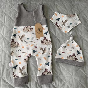 Puede incluir: Un conjunto de bebé de tres piezas con un pelele gris y blanco, un babero y un gorro anudado. La tela blanca está estampada con un castillo, búhos, gafas y otros elementos mágicos. El pelele y el gorro tienen ribetes grises.