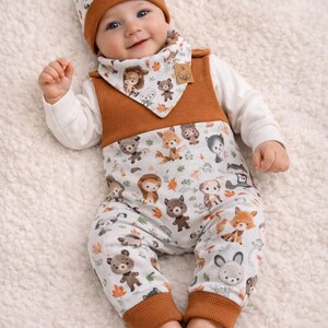 Puede incluir: Un bebé con un conjunto de ropa a juego con un tema de animales del bosque. El conjunto incluye un gorro, un babero y un mono. El mono presenta un fondo azul claro con varias ilustraciones de animales del bosque y puños marrones. El babero y el gorro tienen un ribete marrón.