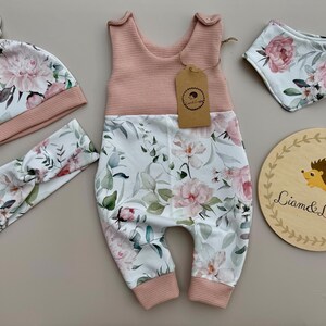 May include: A baby clothing set featuring a pink and white floral print. The set includes a romper, hat, headband, and bib. A wooden sign with a hedgehog illustration and the text "Liam & Luppy" is also included.