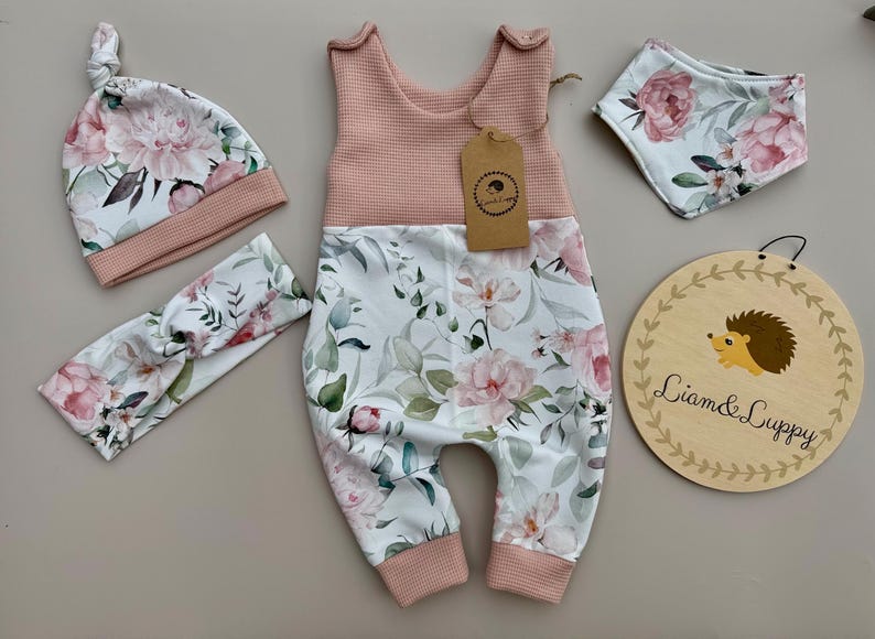 May include: A baby clothing set featuring a pink and white floral print. The set includes a romper, hat, headband, and bib. A wooden sign with a hedgehog illustration and the text "Liam & Luppy" is also included.