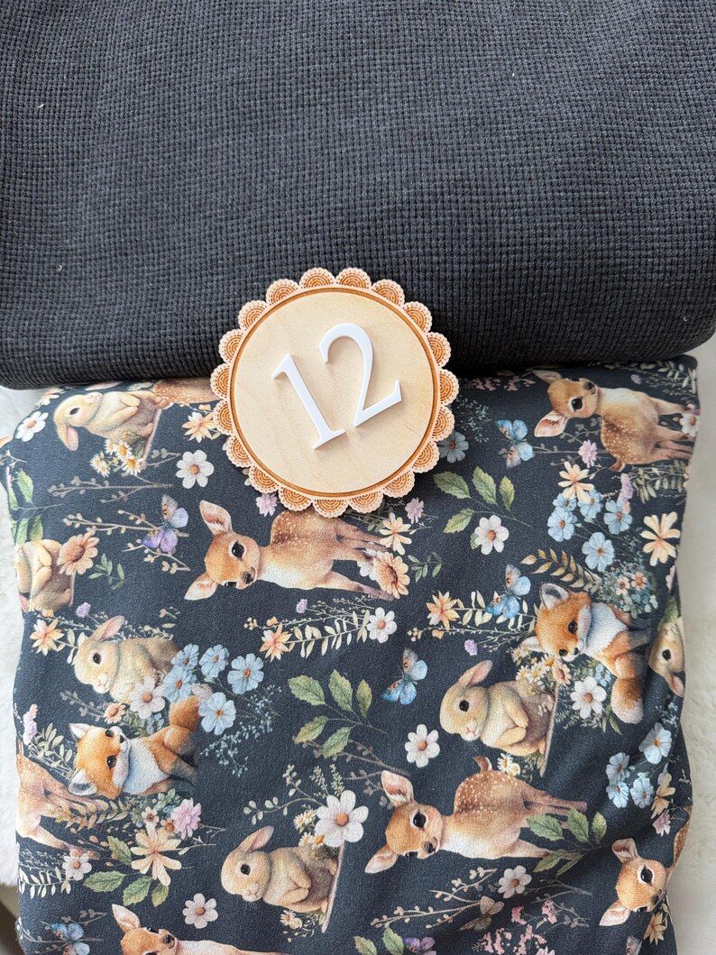Puede incluir: Un c&iacute;rculo de madera con el n&uacute;mero 12 y un borde festoneado descansa sobre una tela gris oscuro y una tela con un estampado de conejos y flores. La tela de conejos tiene un fondo oscuro con conejos, flores y mariposas.