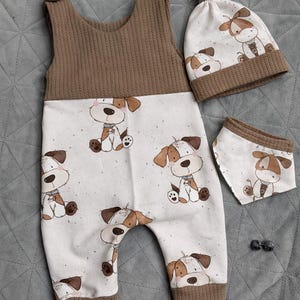 May include: A baby outfit set featuring a brown and white color scheme. The set includes a one-piece jumpsuit, a beanie hat, and a bib, all adorned with a repeating cartoon dog pattern. The jumpsuit has a brown ribbed top and cuffs.