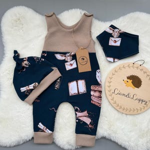 Puede incluir: Un conjunto de ropa de bebé con un mono azul marino, gorro y babero a juego con temática mágica. El conjunto incluye una parte superior y ribete de punto de gofre beige, y un letrero de madera con una ilustración de erizo y el texto "Liam & Luppy."