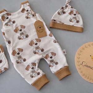 May include: A baby outfit set featuring a white romper, bib, and hat, all adorned with a repeating pattern of brown and white puppy illustrations. The set includes a wooden sign with a hedgehog illustration and the text "Liam & Luppy."