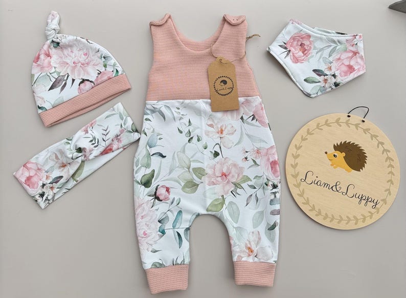 May include: A baby clothing set featuring a pink and white floral print. The set includes a romper, hat, headband, and bib. A wooden sign with a hedgehog illustration and the text "Liam & Luppy" is also included.