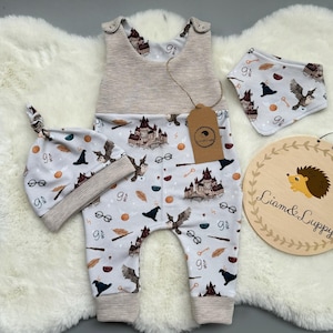 Puede incluir: Un conjunto de ropa de bebé con un pelele, gorro y babero de color gris claro con un estampado repetido de un castillo, gafas y un búho volador. Incluye un letrero de madera con una ilustración de erizo y el texto "Liam & Luppy".