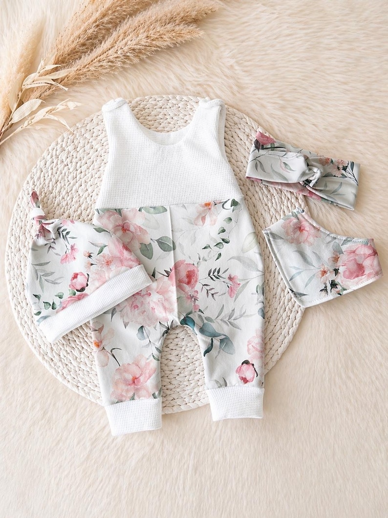 May include: A baby outfit set featuring a white waffle-knit top and floral print pants with pink flowers and green leaves. The set includes a matching hat, bib, and headband. The items are arranged on a woven mat.