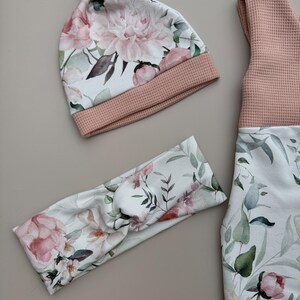 May include: A baby clothing set featuring a floral print with pink peonies and green leaves. The set includes a knotted hat, a headband, and a sleeveless top with matching leggings. The hat and top have a pink waffle knit trim.