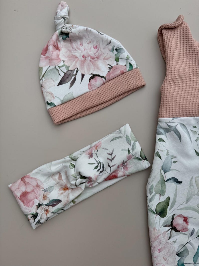 May include: A baby clothing set featuring a floral print with pink peonies and green leaves. The set includes a knotted hat, a headband, and a sleeveless top with matching leggings. The hat and top have a pink waffle knit trim.