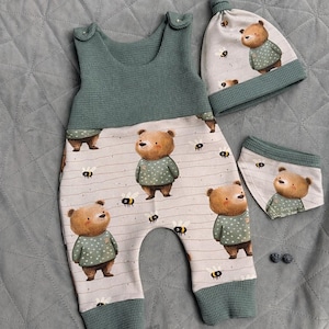 Puede incluir: Un conjunto de ropa de bebé en verde y beige. El conjunto incluye un mono, un gorro y un babero, todos adornados con un patrón repetido de osos y abejas de dibujos animados. El mono tiene una parte superior y puños verdes, con un estampado de oso y abeja en la parte inferior.