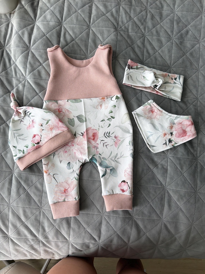 May include: A baby clothing set featuring a pink and white floral print. The set includes a sleeveless romper, a knotted hat, a headband, and a bib. The romper has a pink waffle-knit top and floral print bottoms.