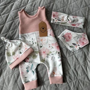 May include: A baby clothing set featuring a pink and white floral print. The set includes a sleeveless romper, a knotted hat, a headband, and a bib. The romper has a pink waffle-knit top and floral print pants.