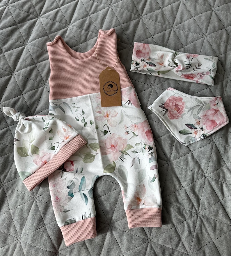 May include: A baby clothing set featuring a pink and white floral print. The set includes a sleeveless romper, a knotted hat, a headband, and a bib. The romper has a pink waffle-knit top and floral print pants.