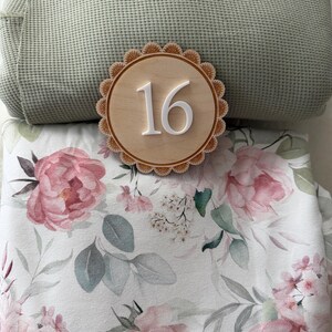 Puede incluir: Un letrero de madera con el n&uacute;mero 16 y un borde festoneado, sobre una manta floral con peon&iacute;as rosas y hojas grises. Una almohada texturizada verde claro est&aacute; en el fondo.