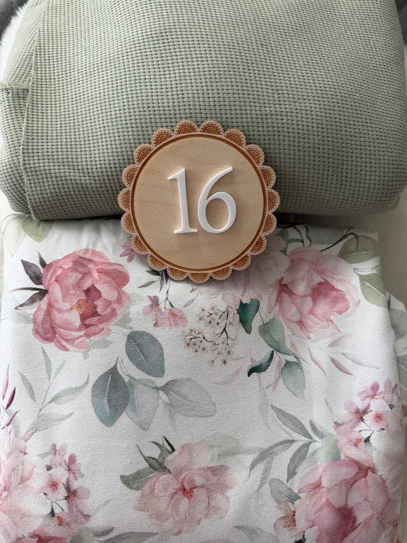 Puede incluir: Un letrero de madera con el n&uacute;mero 16 y un borde festoneado, sobre una manta floral con peon&iacute;as rosas y hojas grises. Una almohada texturizada verde claro est&aacute; en el fondo.