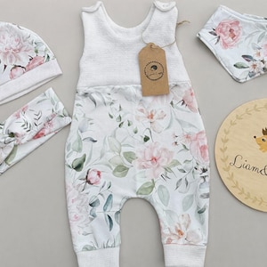 Puede incluir: Un conjunto de ropa de bebé con un estampado floral de flores rosas y blancas y hojas verdes. El conjunto incluye un gorro, una diadema, un babero y un mono. También se incluye un letrero de madera con una ilustración de erizo.
